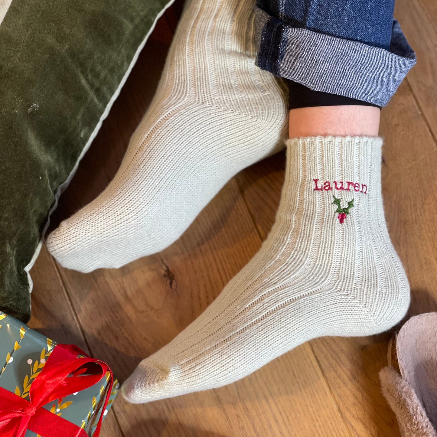 Person wearing white socks with 'Lauren' and a Christmas holly design, sitting on a wooden floor next to a gift box.